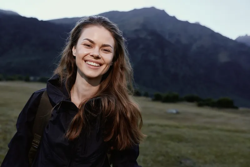 A smiling young woman stands outdoors in a mountainous landscape, wearing a dark jacket and backpack. She radiates confidence and joy, enjoying a bright moment in nature.