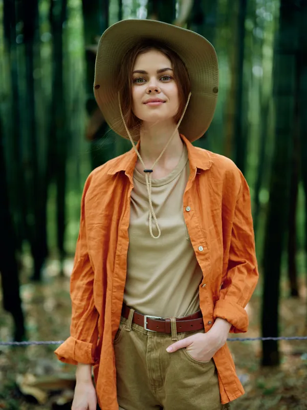 Woman in hat portrait in a bamboo forest outdoors and nature, casual orange shirt with hand in pocket, confident smile, travel and adventure vibe, eco friendly lifestyle.
