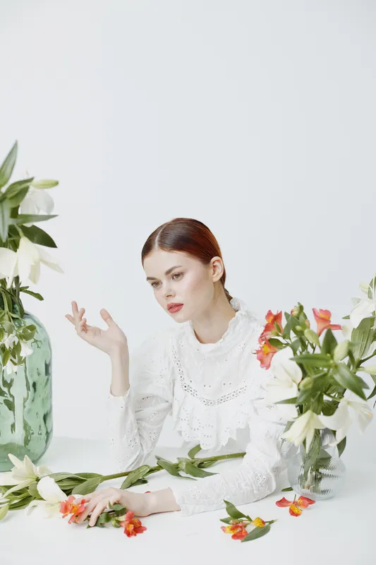 Elegant young woman in white dress surrounded by flowers, conveying beauty and serenity in a bright, minimalist setting with a soft color palette