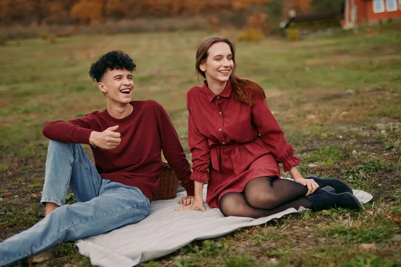 Two friends sit on a blanket outdoors, sharing a candid moment with genuine smiles and relaxed posture, capturing authenticity in everyday connection under soft light.