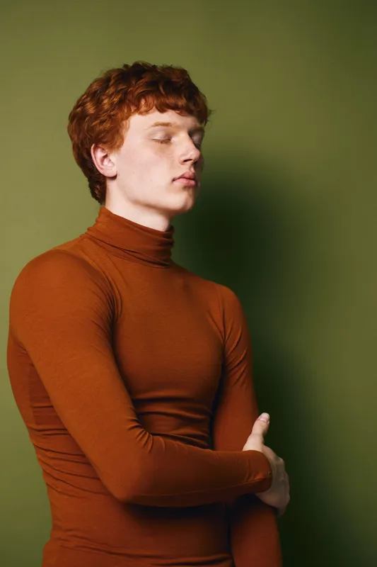 Young man with red hair and freckles wearing a brown turtleneck sweater standing with crossed arms and closed eyes against olive green background. Portrait, calm and thoughtful mood.
