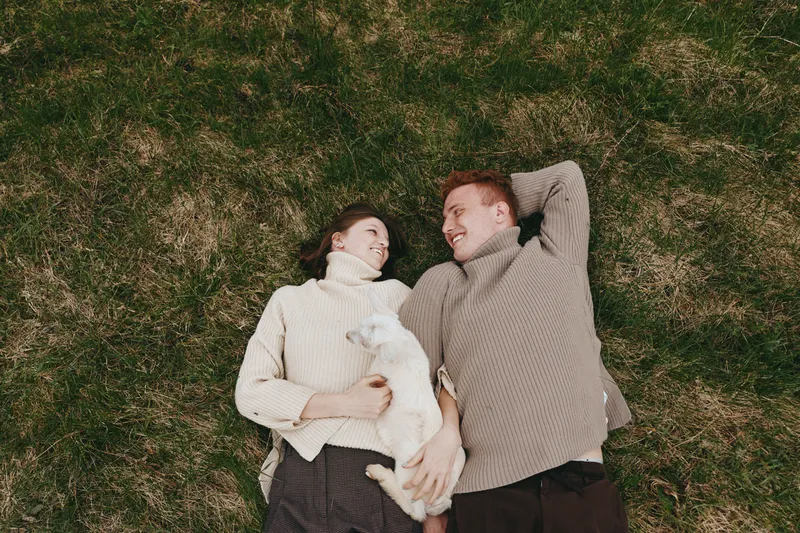 A couple lies on a grassy field, smiling at each other with a small dog tucked between them, cozy sweaters creating a warm, relaxed lifestyle moment.