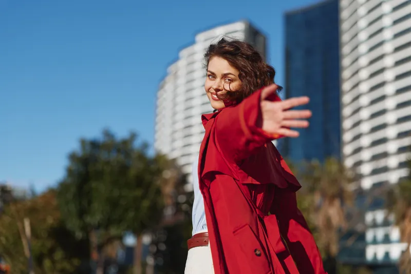 Young woman in a red coat extends her arm toward the camera with a playful smile, standing in a bright urban setting with modern buildings, trees, and blue sky behind.