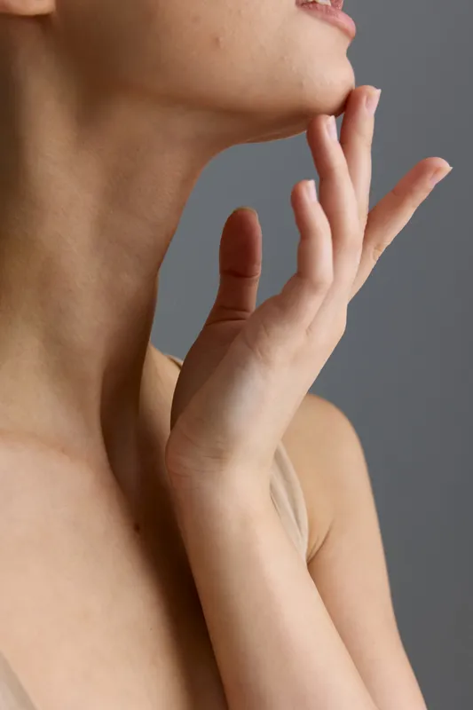 Close-up of woman’s neck and hand with delicate skin care gesture on gray background, natural light highlighting smooth complexion and soft feminine beauty.