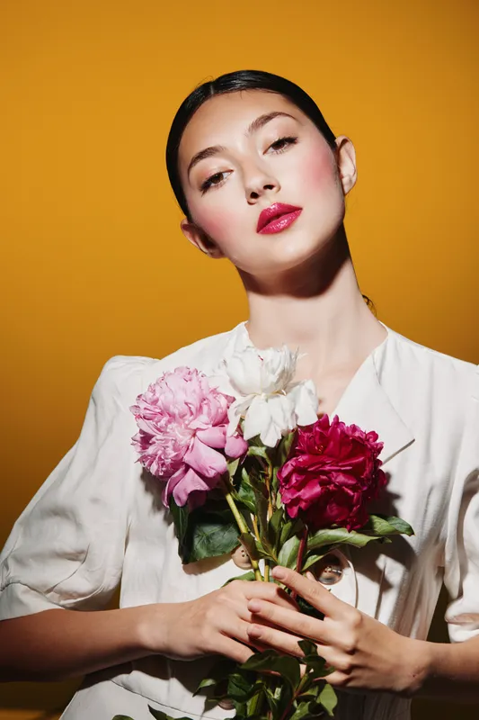 Young woman with vibrant makeup holding bouquet of pink and white flowers against orange background in creative beauty fashion concept with fierce stylish look.