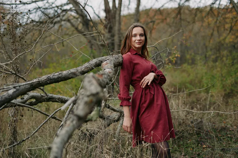 A candid portrait of a woman in a red dress leaning on a fallen tree in an autumn field, natural light and subtle expression conveying authenticity and quiet credibility.
