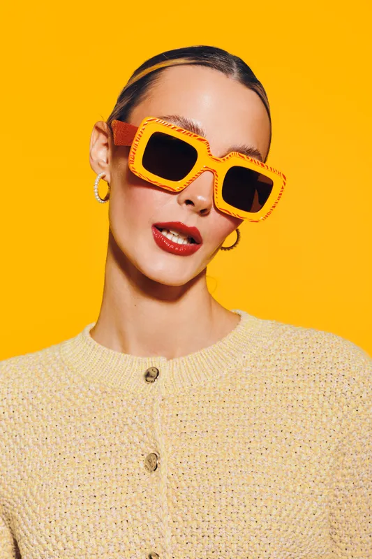 Stylish woman wearing large orange sunglasses and hoop earrings poses against bright yellow background. Fashion portrait with vibrant colors and confident expression.