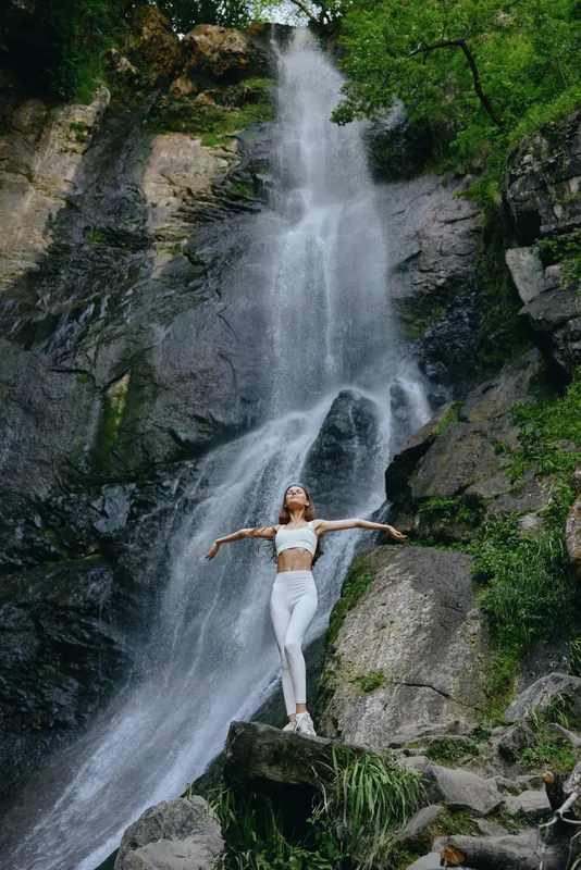 woman standing waterfall arms outstretched nature adventure wearing white outfit surrounded by lush green forest and rocks
