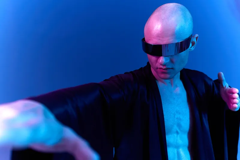 Futuristic man in dark robes poses dramatically with sunglasses, showcasing his unique style. The vibrant blue lighting creates a striking atmosphere.