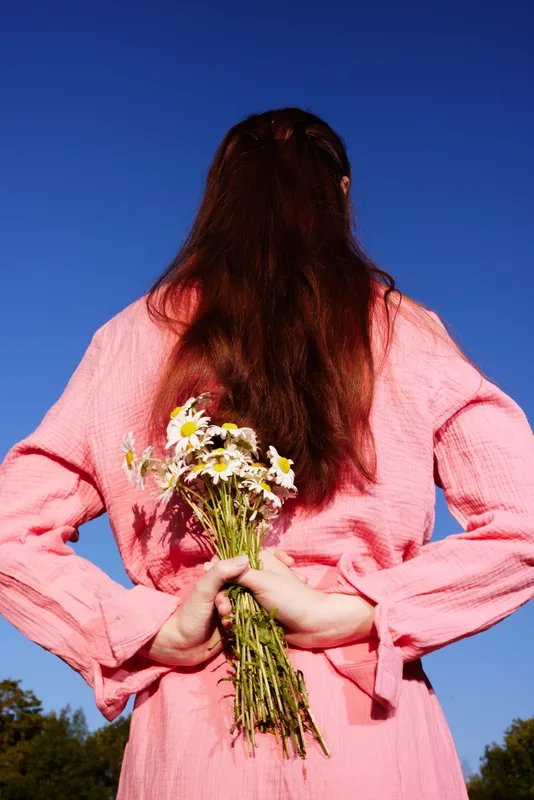 Young model with long hair holding flowers behind her back, wearing a pink outfit against a blue sky, conveying a cheerful and playful spring atmosphere