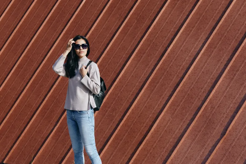 Woman wearing sunglasses with a backpack poses against a diagonal wooden wall, casual denim jeans and a light sweater, street fashion vibe, modern urban backdrop.