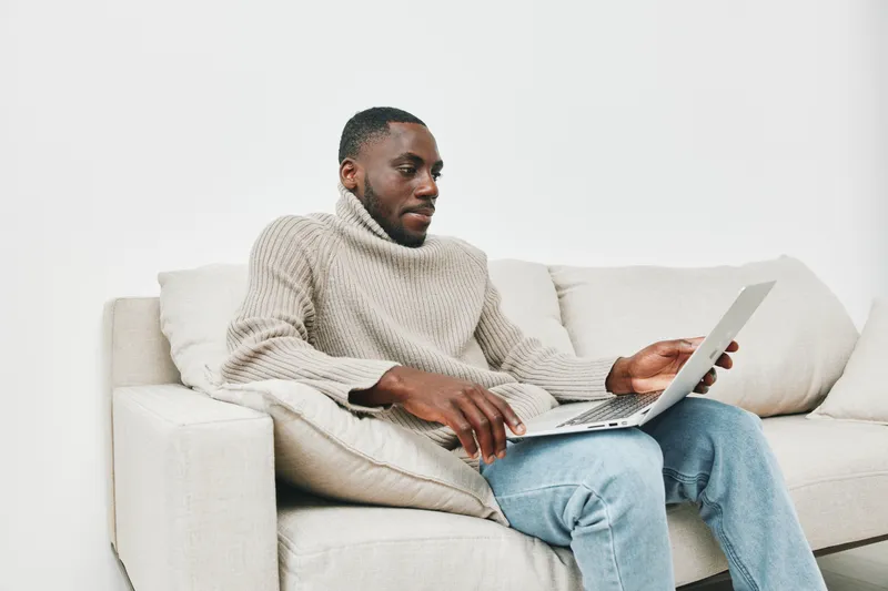 Young African man in cozy sweater working from home on laptop, showcasing productivity and relaxation in a modern living room