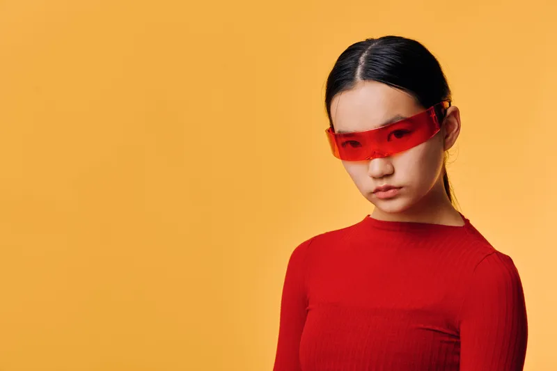 Young woman wearing red futuristic sunglasses and red top, posing with serious face against orange background. Fashion style, portrait, modern look and confident expression in studio.