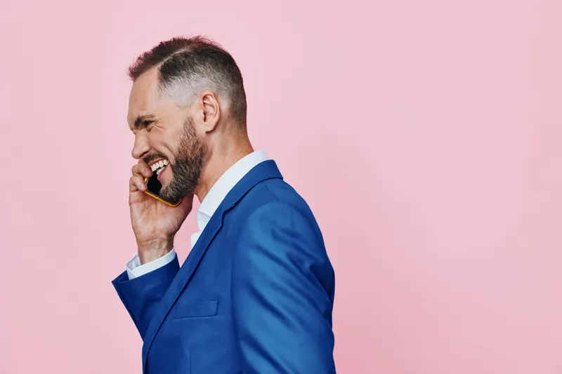 A professional businessman in a sharp blue suit speaks on a mobile, smiling with confident ease against an isolated pink background, conveying effortless communication and leadership.