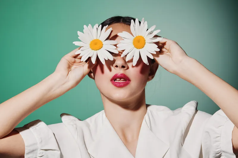 Creative beauty shot of woman holding large white and yellow daisies over eyes on green background, wearing white clothing. Concept of nature, fashion, and floral elegance.