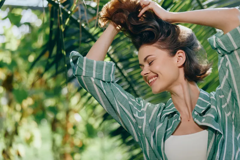 Woman outdoors, smiling joyfully as she adjusts her hair, surrounded by lush greenery, wearing a casual striped shirt, relaxed pose conveying happiness and natural beauty.