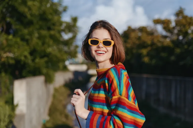 Woman smiling in a rainbow striped sweater and yellow sunglasses, young adult walking outdoors in a sunny suburban park, short brown hair, casual colorful fashion and joyful travel vibe