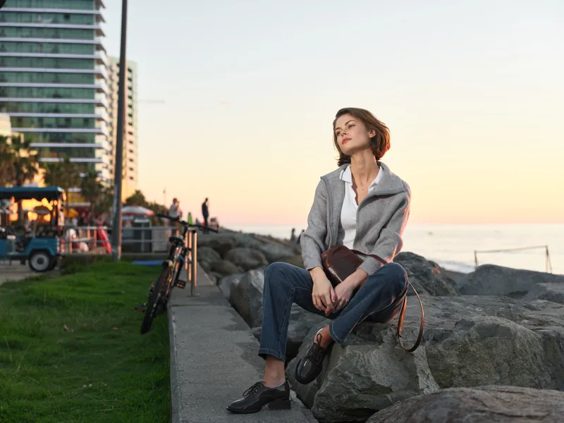 A stylish woman sits on rocky shoreline at sunset, wearing a blazer and jeans. She gazes upward with a calm, thoughtful expression, creating a warm, contemporary mood.