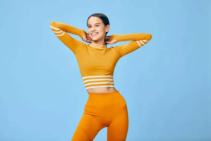 Young woman in orange activewear stretching with hands behind her head, smiling confidently in a studio sports portrait against a clean blue background.