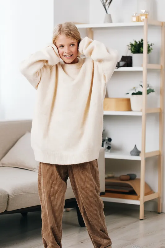 Young girl wearing an oversized cream sweater and corduroy pants poses playfully indoors, hands raised to her head, smiling in a cozy living space with soft natural light.