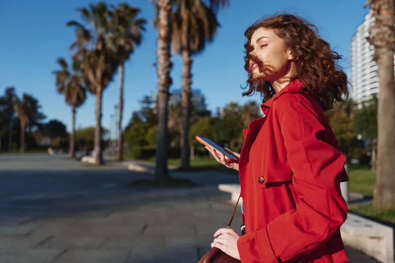Young woman in red coat holds smartphone walks outdoors on sunny day with palm trees in background enjoys fresh air and casual urban lifestyle.