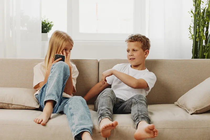 Two children sitting on beige sofa playfully interacting, casual clothing and barefoot in bright living room with natural light and plants in background.