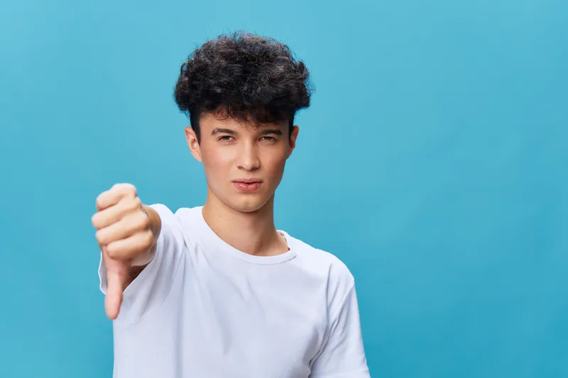 Young man with curly hair showing thumb down gesture against blue background, expressing disapproval, casual white t-shirt, isolated studio portrait.