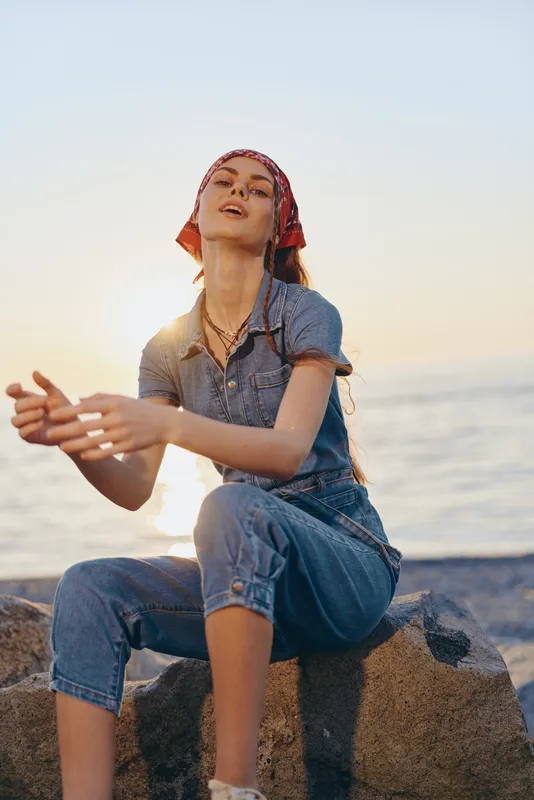 lifestyle woman in modern boho-western denim street style sitting by the sea at sunset with relaxed pose and film color tones