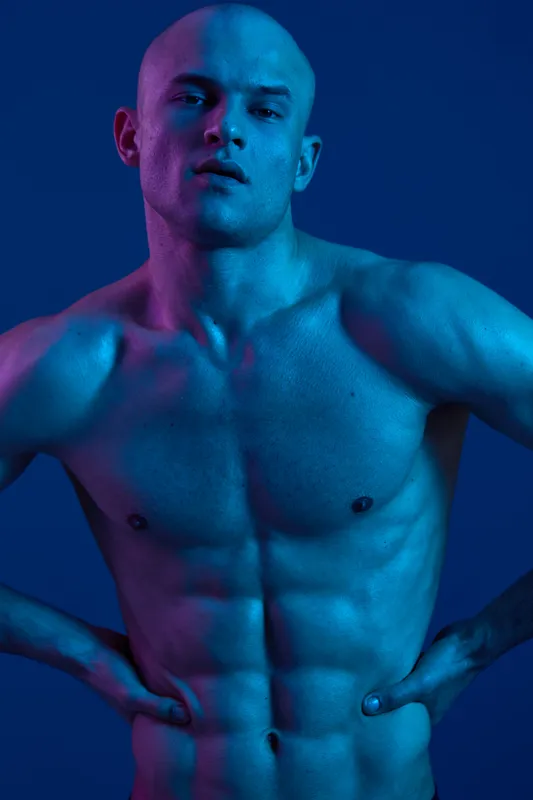 Handsome man with a fit physique poses dramatically against a dark blue background, showcasing artistic lighting and shadows that enhance his muscular structure.
