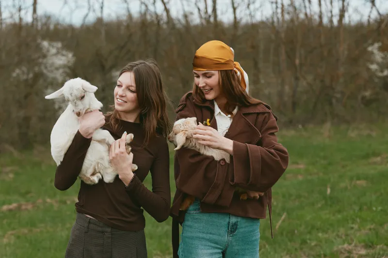 Cheerful women outdoors, holding a kid goat and a young goat, enjoying playful interaction, warm coats, soft breeze, natural colors, rural setting, candid moment, sunlight on faces, wholesome