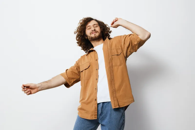 Happy young man with curly hair dancing, wearing a casual brown shirt and white t shirt, showcasing joy and freedom against a minimalist white background