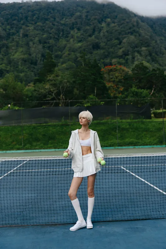 Young female tennis player in sporty white outfit, holding tennis balls on outdoor court, surrounded by lush greenery, exuding confidence and athleticism