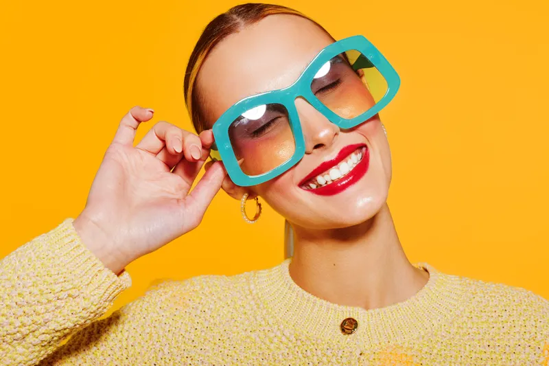Smiling woman with bright red lipstick wearing oversized blue sunglasses and yellow sweater poses against vibrant orange background. Concept of fashion, summer style, and joyful mood.