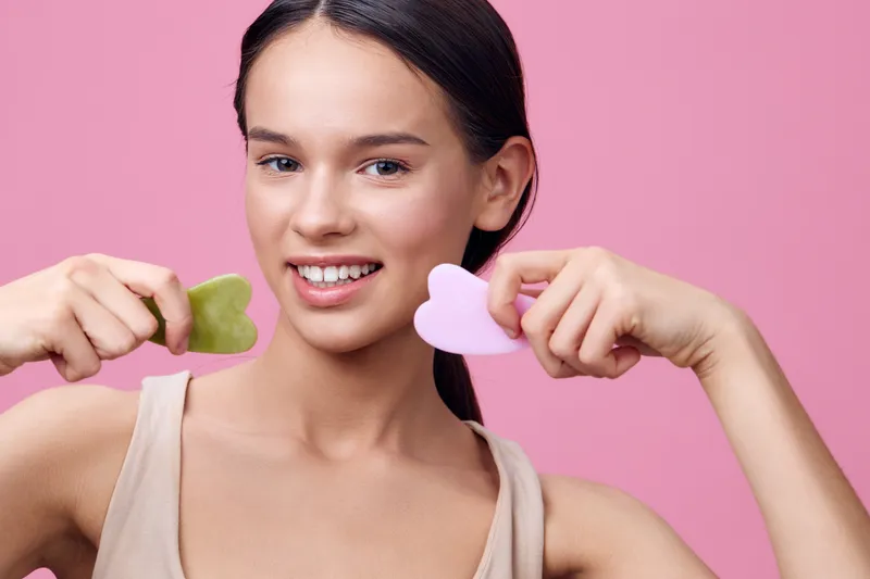 Young woman holding facial tools, smiling with a vibrant pink background, showcasing healthy skincare concepts and self care emotions