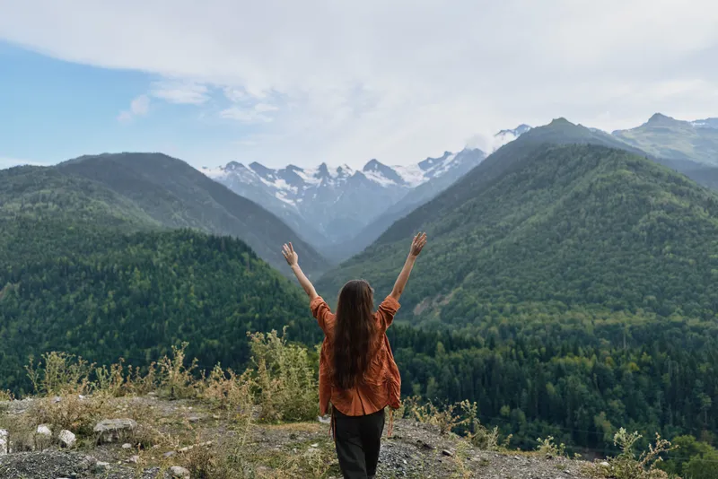 Woman stands on a rocky overlook with arms raised, embracing a vast mountain landscape, clear sky, evergreen forest, and distant peaks that spark adventure and freedom