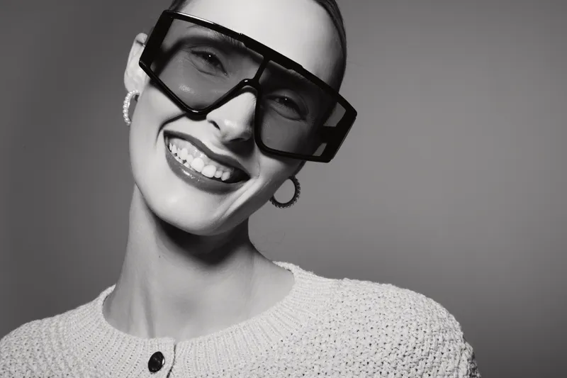 Smiling caucasian woman with light skin wearing oversized glasses and knitted sweater poses happily in monochrome studio portrait with stylish earrings.