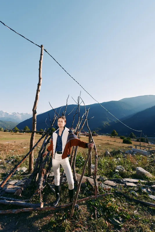 Person portrait in countryside mountains by a rustic fence, fashion model posing in a nature field, stylish jacket and boots, scenic rural landscape with clear blue sky and meadow.