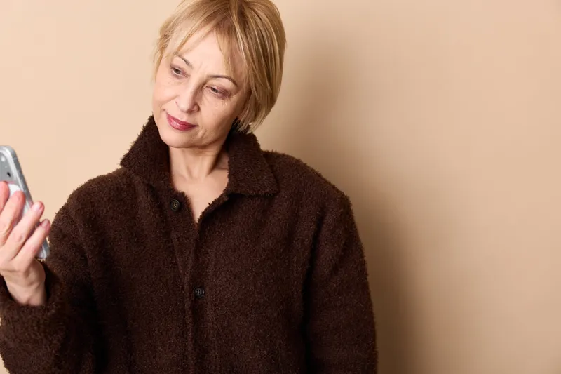 Senior woman with short blond hair holding a smartphone, communication portrait, warm beige background, soft lighting, smile, technology at home, relaxed mood