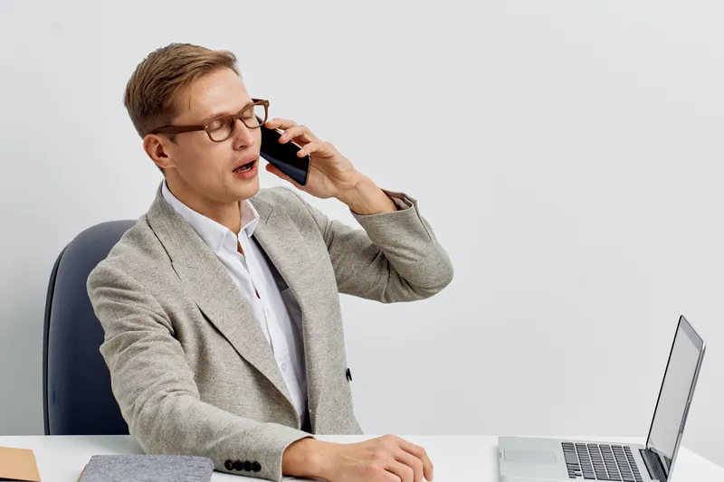 Businessman talking on mobile phone indoors with laptop in office, casual formal attire, middle-aged, serious expression, modern workspace, neutral background, professional communication concept