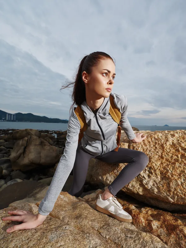 Athletic model climbing a rocky terrain by the sea, showcasing determination with a focused expression, wearing a gray sports jacket and athletic shoes