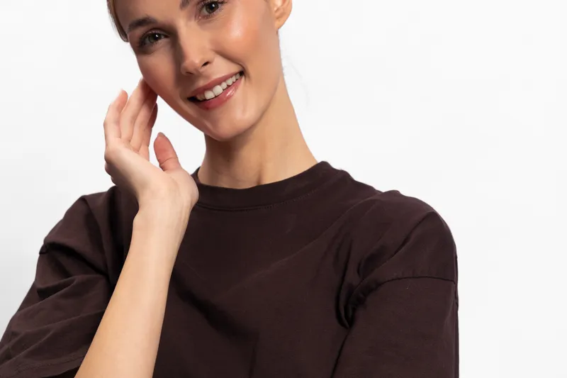 Smiling woman with clear skin and short hair touches her face gently in a brown sweater against a white background. Concept of beauty, skincare, and natural confidence.