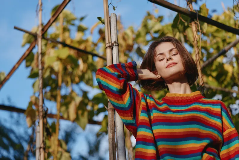 Woman in a colorful striped sweater leaning against a wooden trellis outdoors, young adult with short bob, smiling peacefully in warm sunlight for casual lifestyle and travel vibe.
