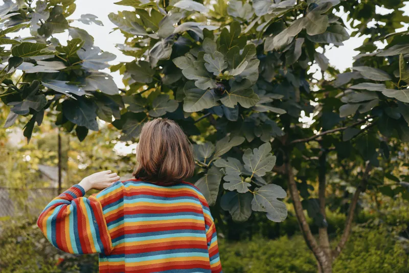Woman in striped sweater seen from back, short brown hair touching hair, casual outfit in green garden, relaxed mood, leaning under tree for lifestyle travel or outdoor fashion portrait.