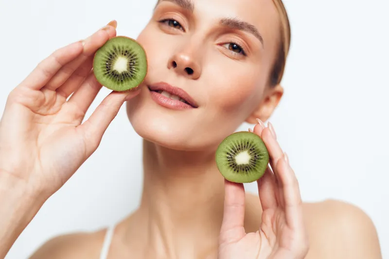 Young woman with clear skin holding two halved kiwi fruits near her face on a white background. Concept of healthy lifestyle, natural beauty, skincare, and wellness.