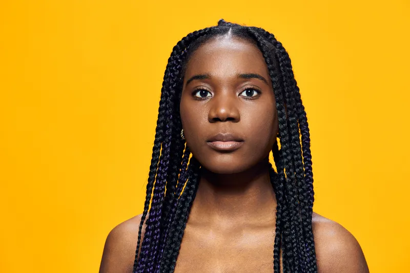 A portrait of a young African girl with braids against a vibrant yellow background, expressing confidence and self assurance through her intense gaze
