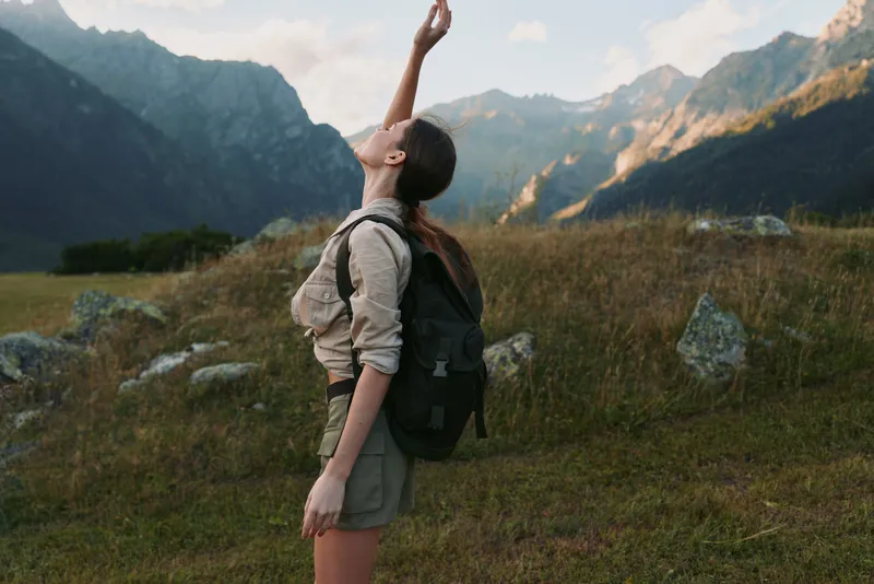 Young woman with backpack hiking in mountains enjoys nature and fresh air, stands on grassy field surrounded by rocks and peaks under cloudy sky.