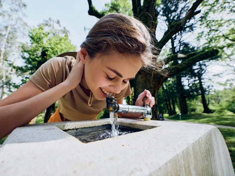 Girl drinking water from a fountain in a sunny park, smiling and leaning over the basin to sip the fresh stream. Outdoor child enjoying playful hydration and summer joy.