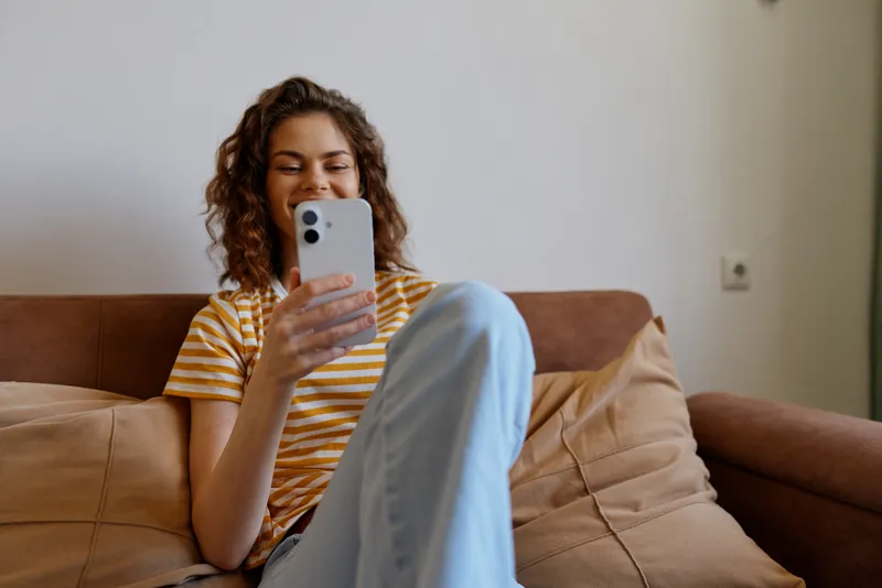 Young woman sits on a sofa at home, wearing a striped shirt, using a smartphone, relaxed mood, cozy living room setting, natural light, candid lifestyle portrait with casual atmosphere.