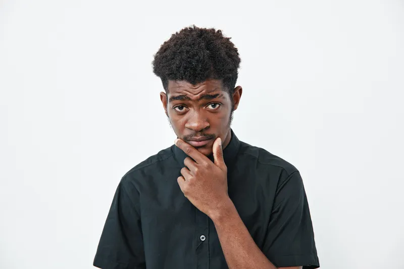 Young black man with dark skin and curly hair wearing black shirt looking thoughtful with hand on chin isolated on white background. Portrait of pensive african male.