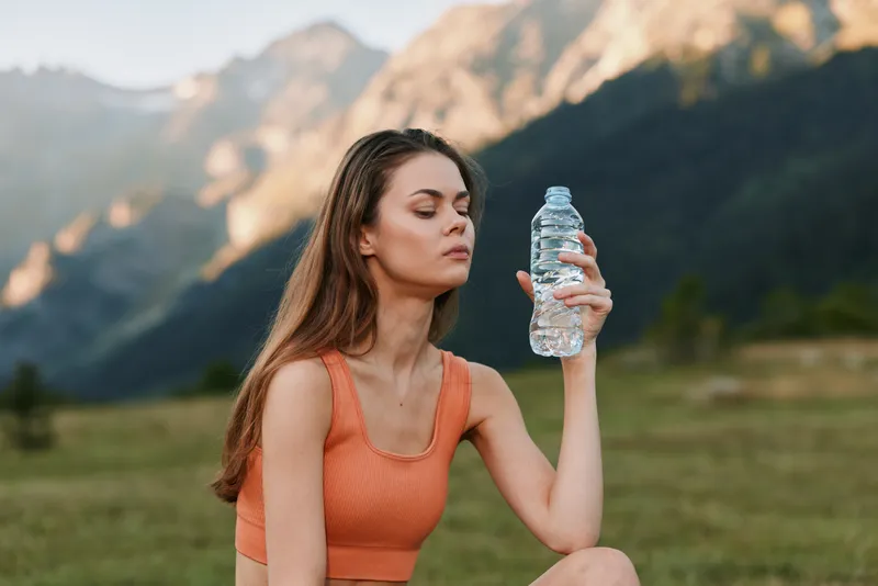 A focused young woman sits outdoors in a scenic valley, wearing an orange athletic top and holding a water bottle as she rests, reflecting calm and fitness in nature.